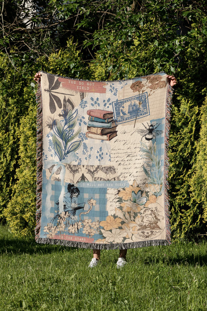 A person is holding up a colorful blanket with various illustrations and text, including a book, flowers, and a bee, in a grassy area surrounded by trees.