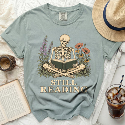 Skeleton reading a book surrounded by flowers with text "STILL READING".