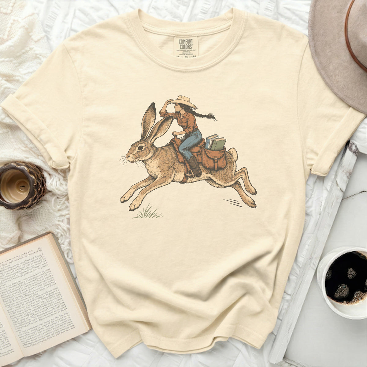 Cream t-shirt with a drawing of a cowgirl riding a rabbit, carrying books.
