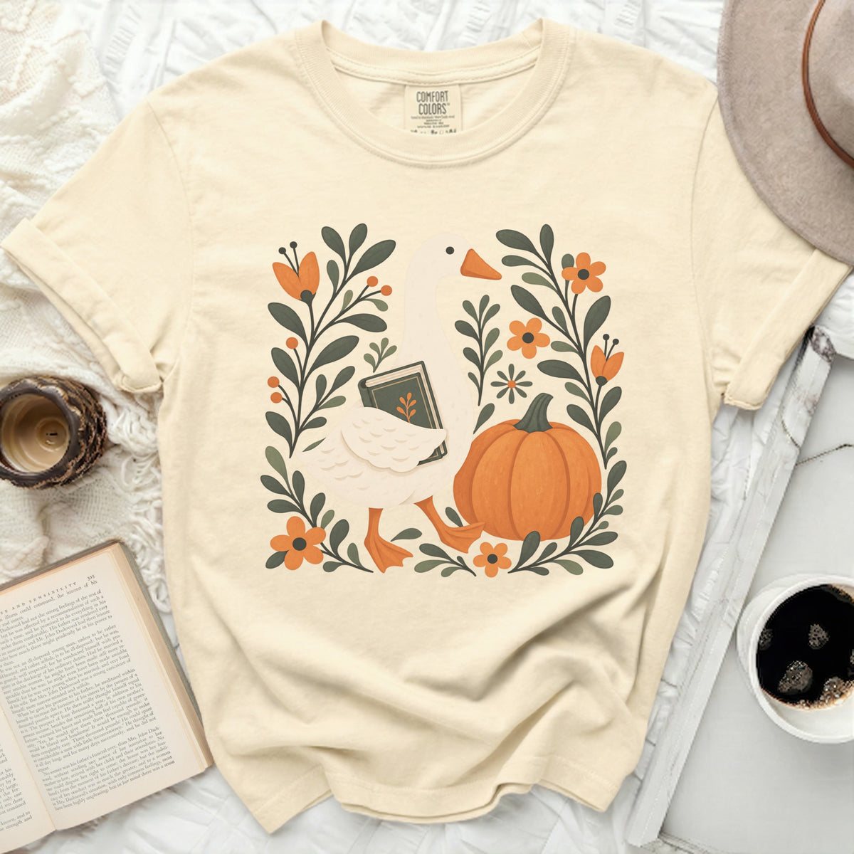 Cream t-shirt with white goose holding book, pumpkin, and floral design.