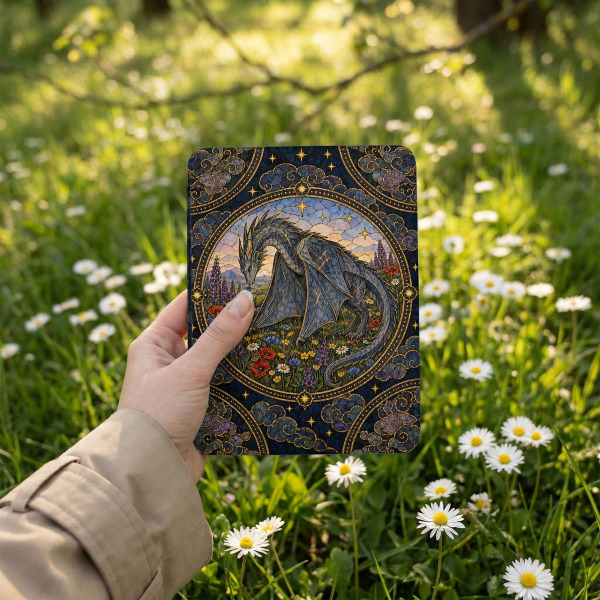 A hand holds an e-reader featuring an intricate fantasy dragon illustration against a daisy-filled meadow background.