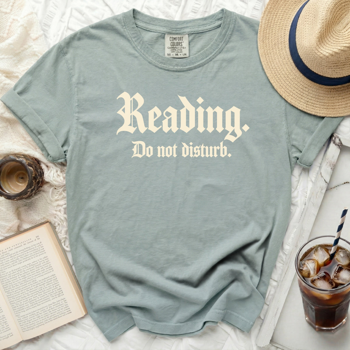 Sage green t-shirt featuring the white text Reading. Do not disturb, surrounded by a straw hat, book, and iced drink.