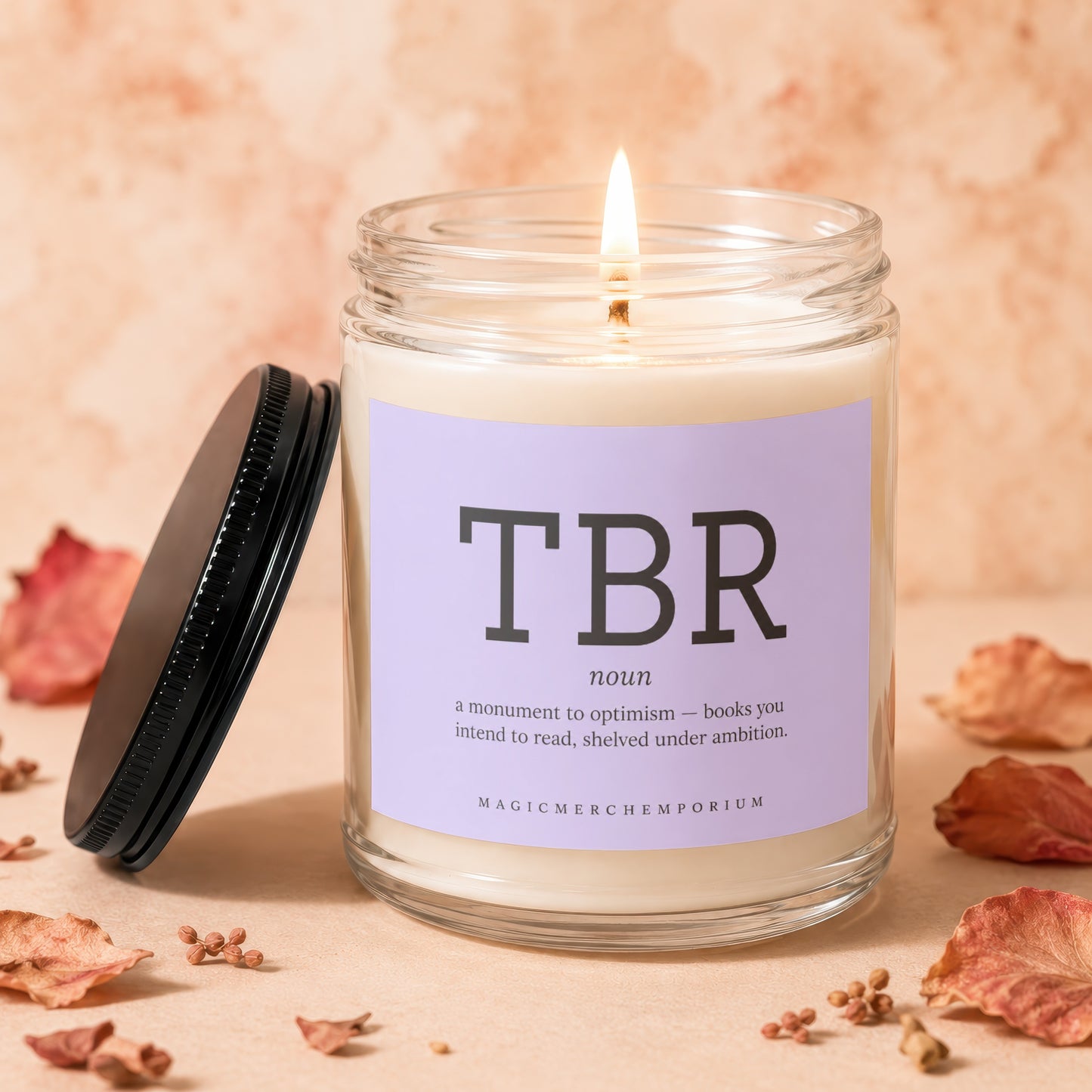 Candle with "TBR" definition, lid off, lit flame, surrounded by dried leaves.