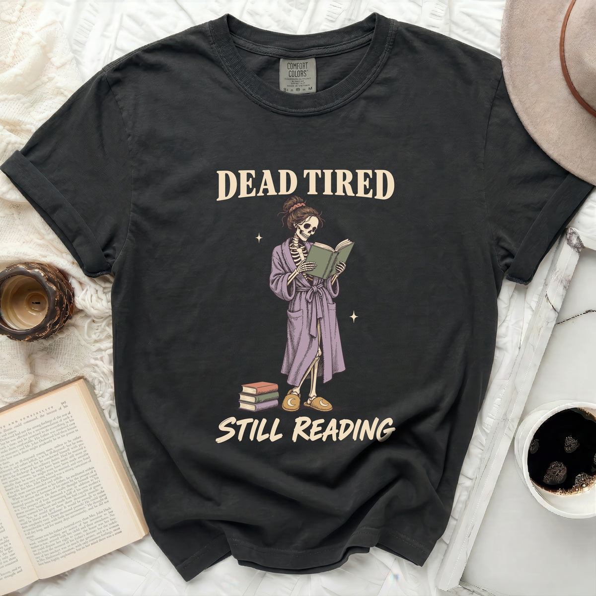 T-shirt with skeleton graphic reading a book, text says "DEAD TIRED STILL READING".