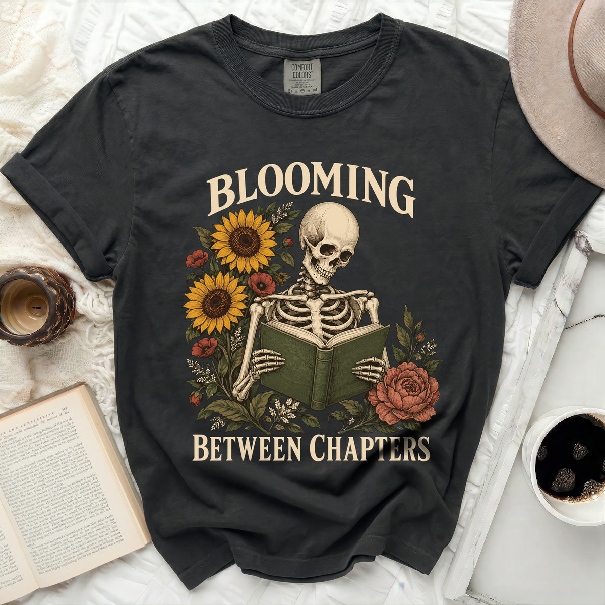 A charcoal t-shirt features a skeleton reading a book surrounded by sunflowers and the text Blooming Between Chapters.