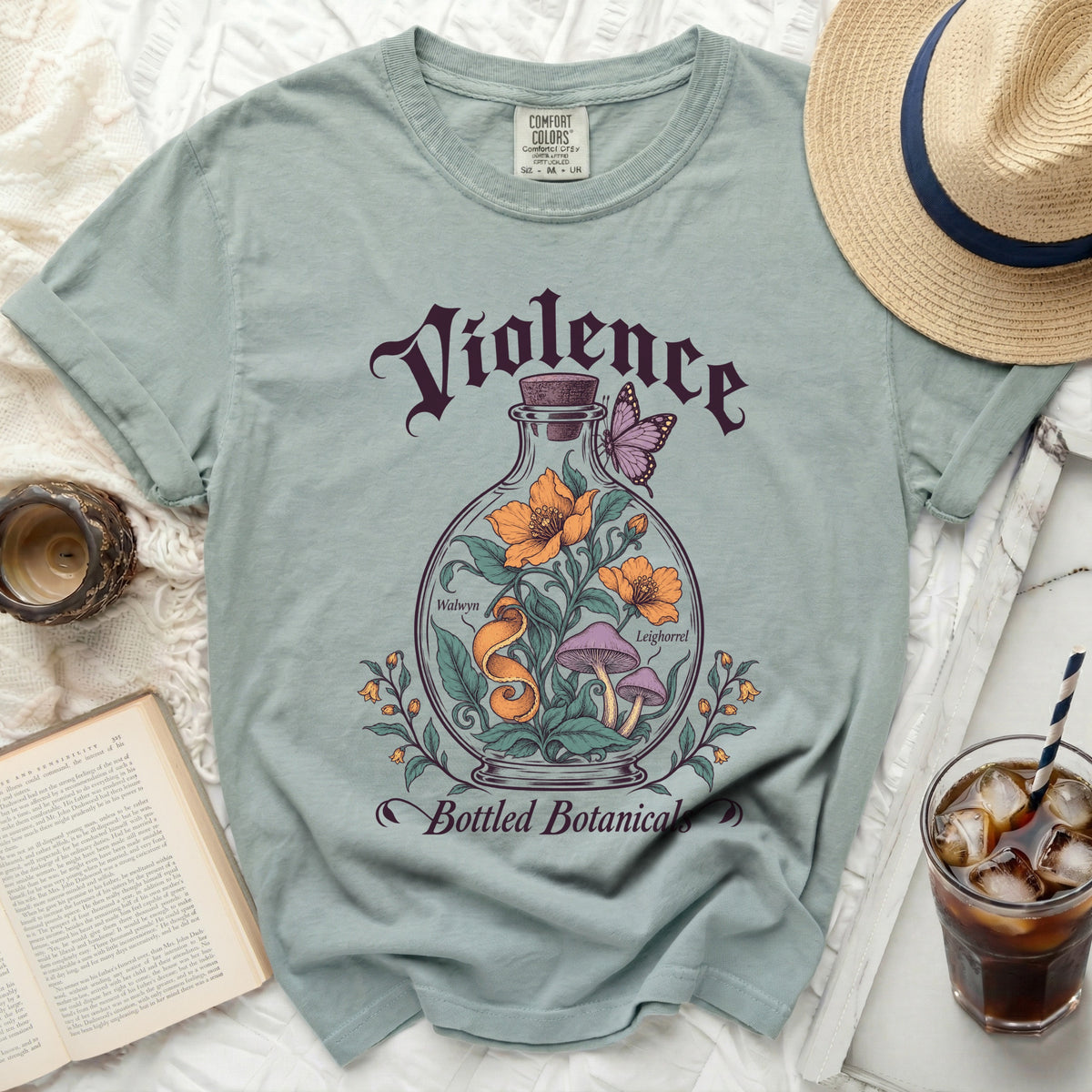 Sage green t-shirt with a graphic of a glass bottle containing flowers, mushrooms, and a butterfly, surrounded by text reading "Violence Bottled Botanicals".
