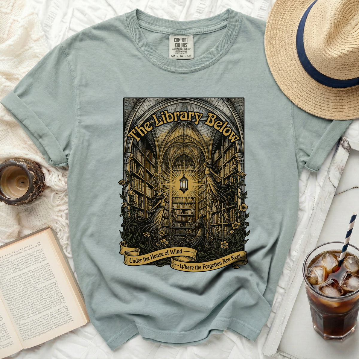 Light blue t-shirt with "The Library Below - Under the House of Wind - Where the Forgotten Are Kept" graphic.