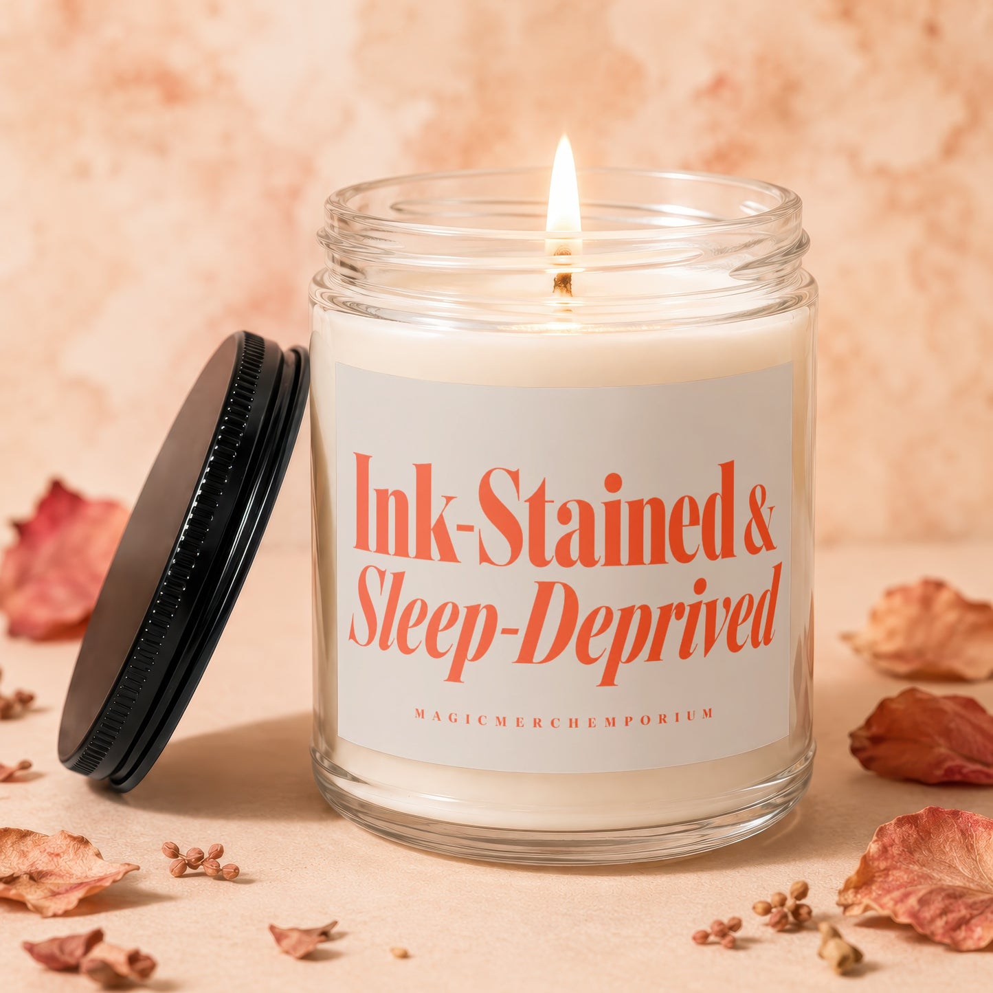 Candle with "Ink-Stained & Sleep-Deprived" text, lid off, lit wick, dried leaves and seeds.