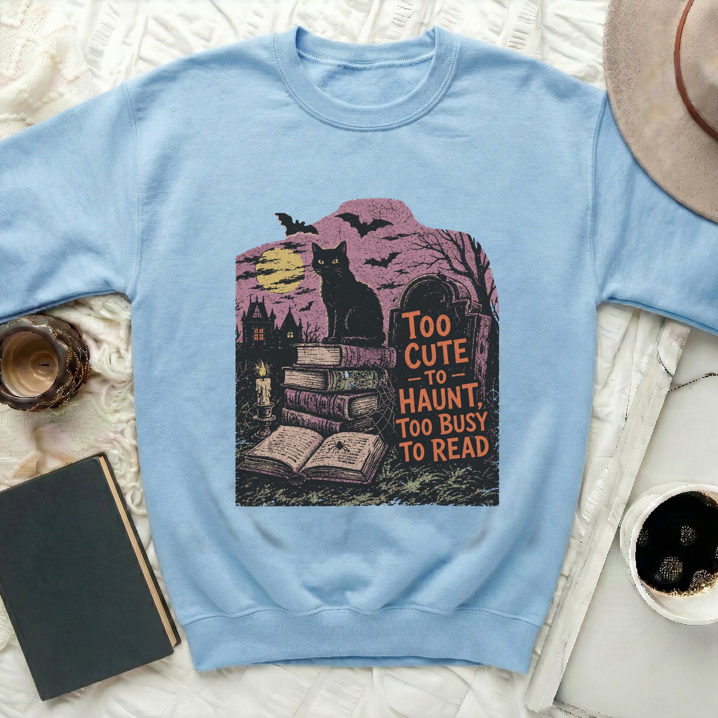 Light blue sweatshirt with graphic of black cat, books, graveyard, and "Too Cute to Haunt" text.