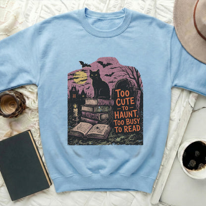 Light blue sweatshirt with graphic of black cat, books, graveyard, and "Too Cute to Haunt" text.