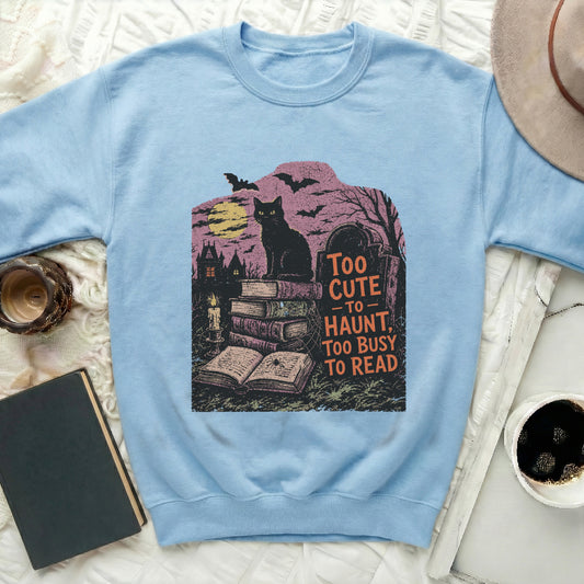 Light blue sweatshirt with graphic of black cat, books, graveyard, and "Too Cute to Haunt" text.