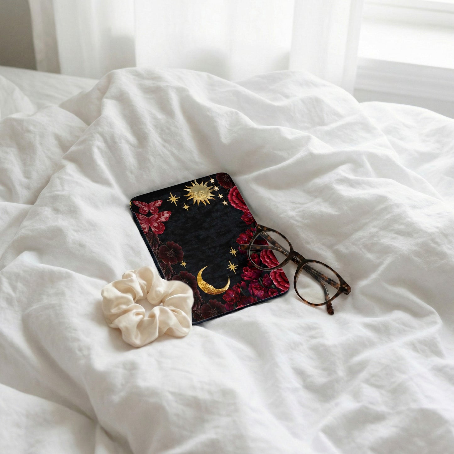 Case with celestial and floral design, scrunchie, and glasses on white bedding.