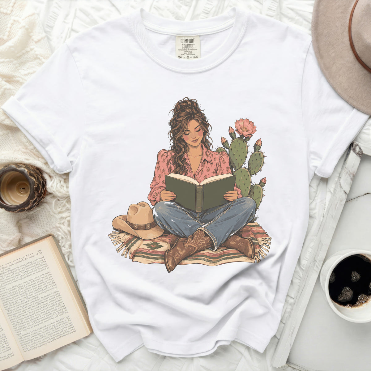 White t-shirt with illustration of woman reading by cactus, wearing cowboy boots.