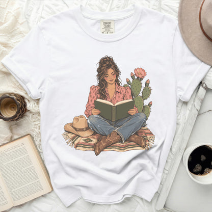 White t-shirt with illustration of woman reading by cactus, wearing cowboy boots.