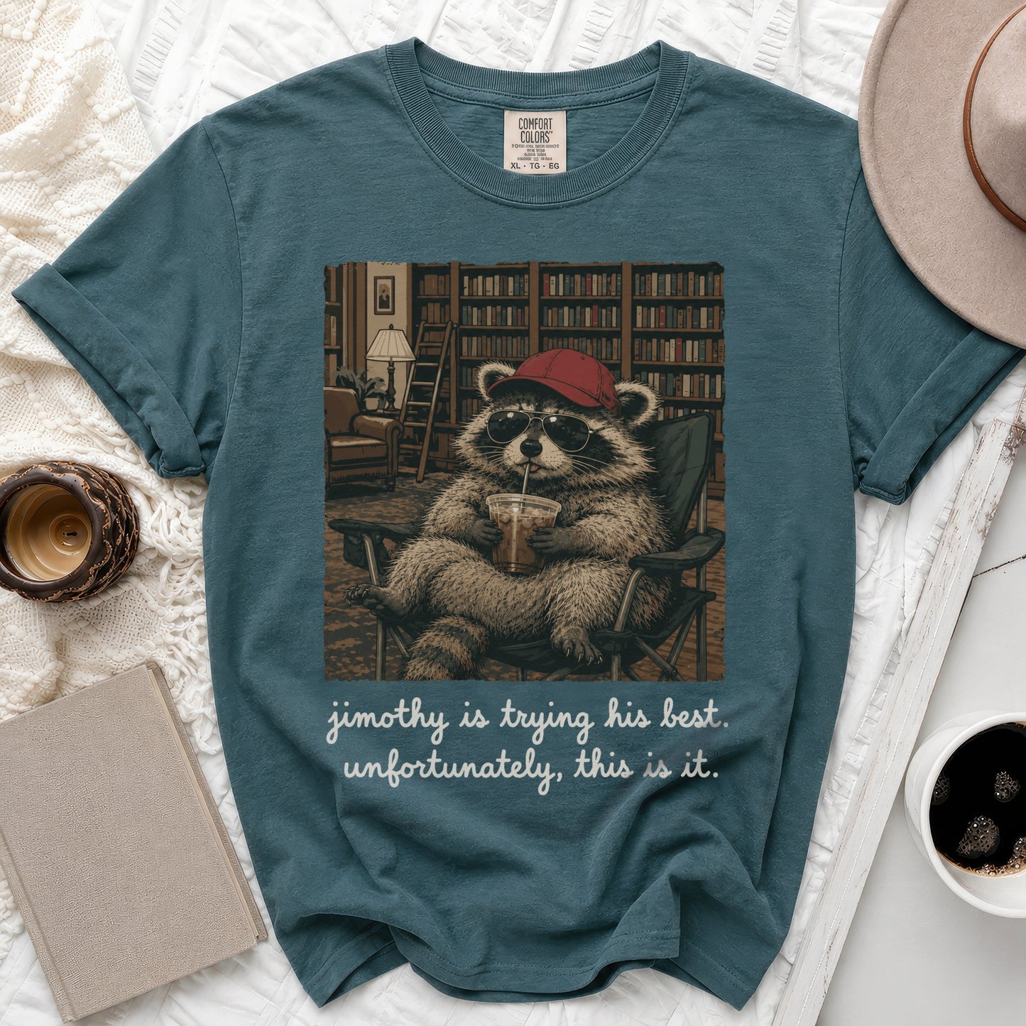 Graphic tee with raccoon in sunglasses and hat holding drink, text "jimothy is trying his best. unfortunately, this is it."