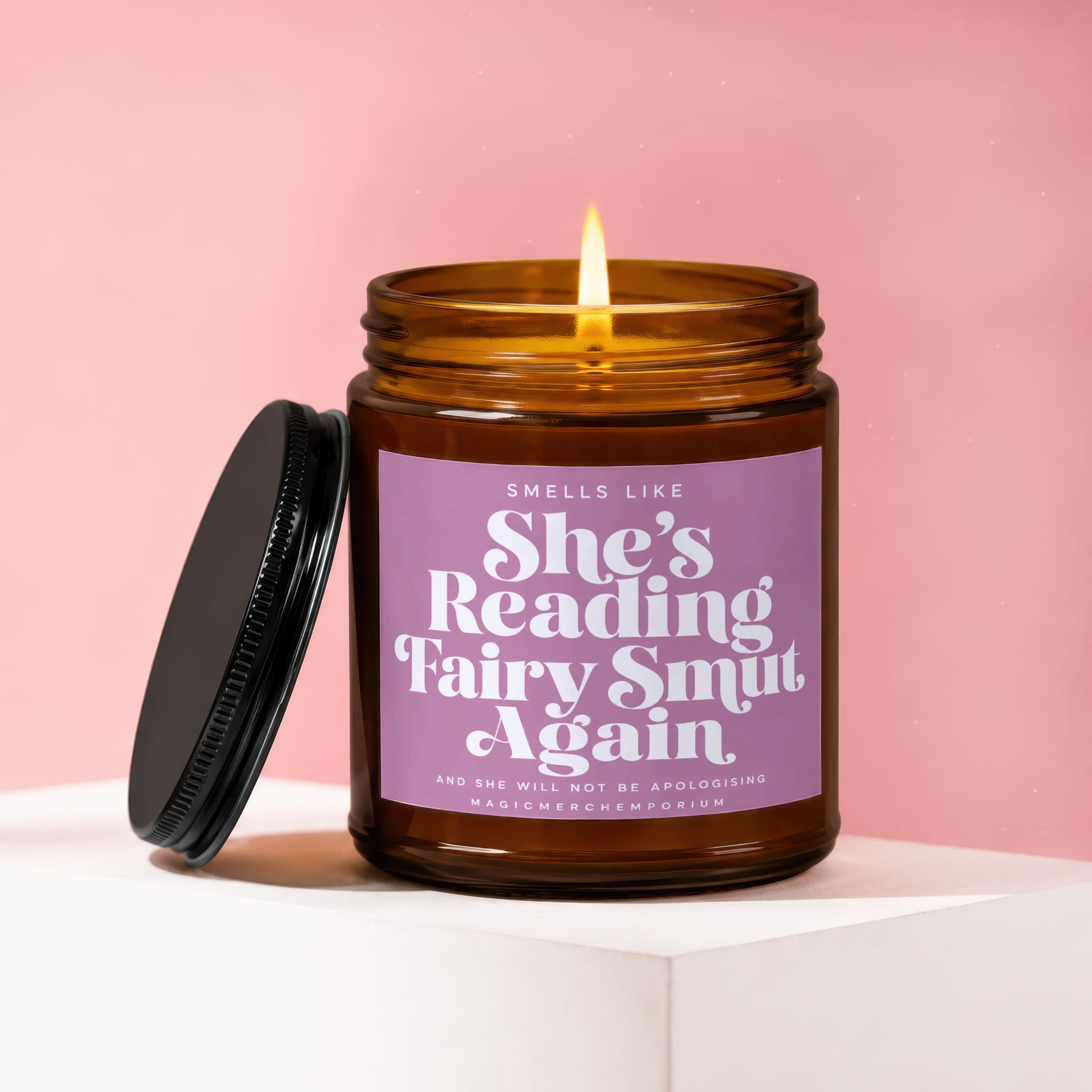 Amber glass candle burning with a purple label reading "Smells Like She's Reading Fairy Smut Again".