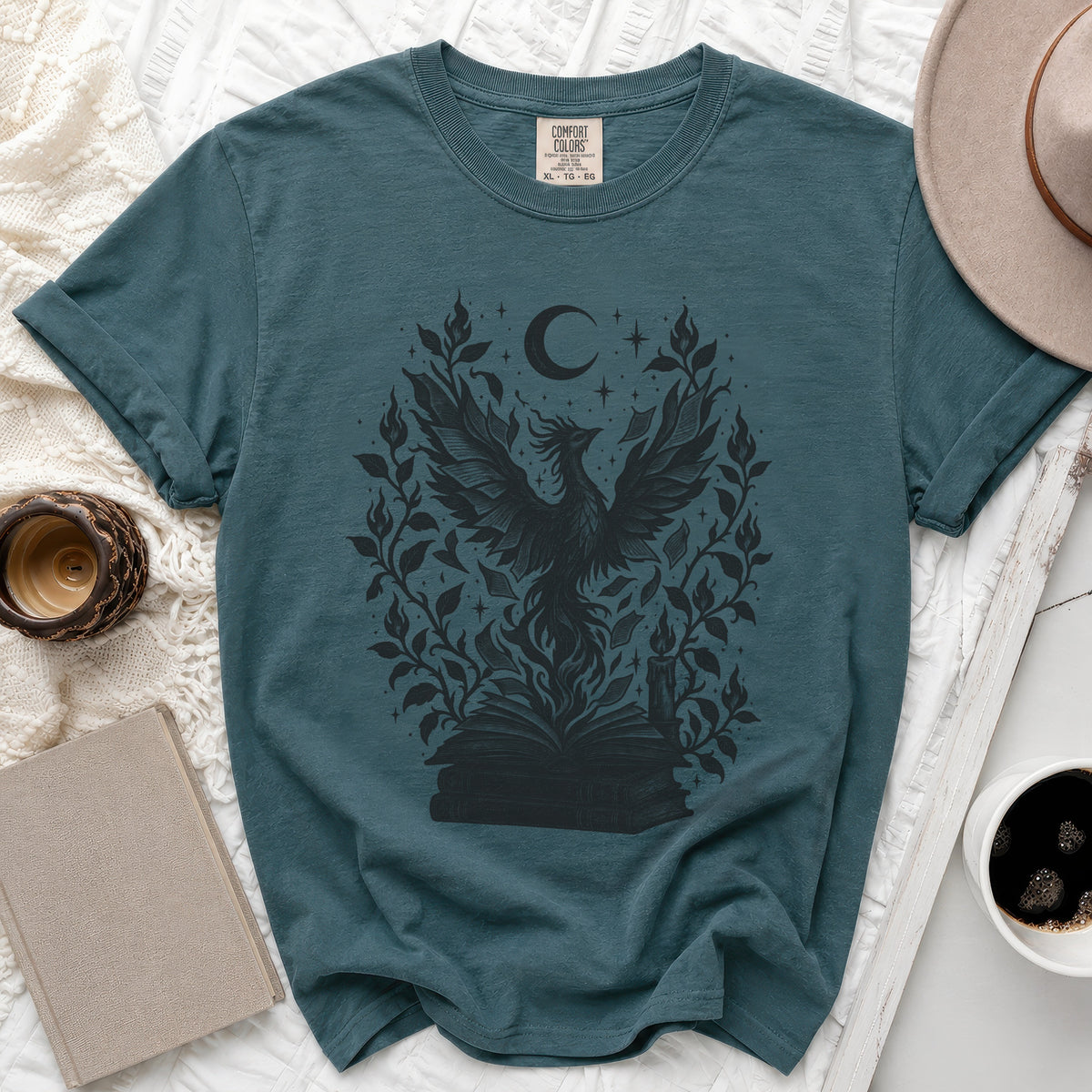 A teal t-shirt featuring a black graphic of a phoenix rising from books surrounded by leaves, stars, and a crescent moon.