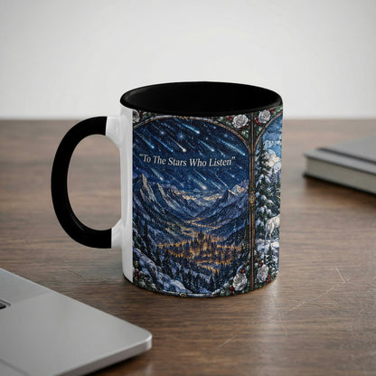 A black-rimmed white ceramic mug sits on a wooden desk, featuring a stained glass-style mountain landscape and the text To The Stars Who Listen.
