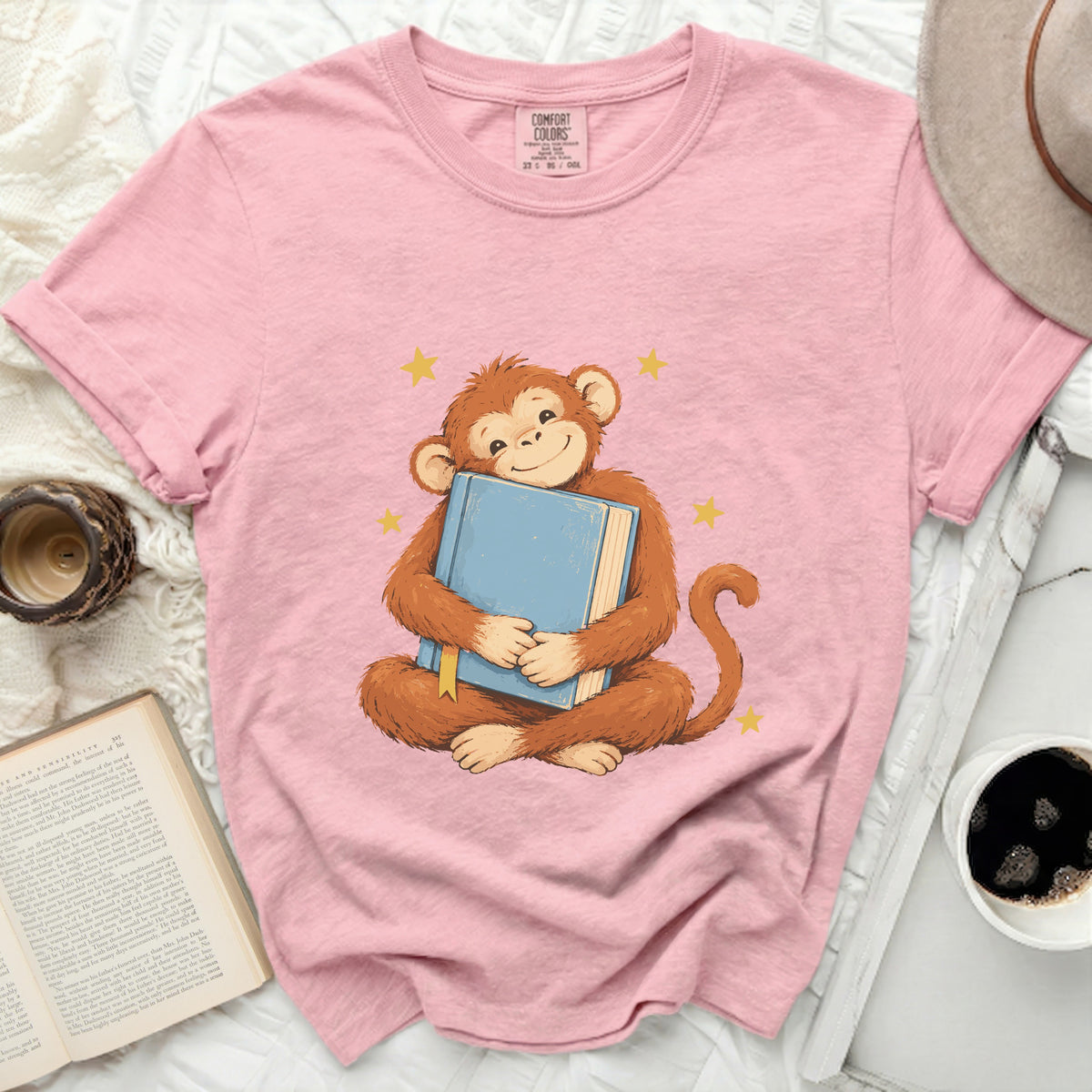 Pink t-shirt with a graphic of a monkey hugging a blue book and yellow stars.
