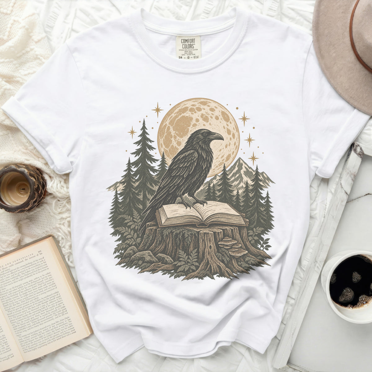 White t-shirt with a graphic of a raven on a book in front of a moon and forest.