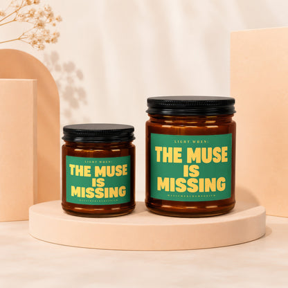 Two amber glass jars with black lids, candles inside, labeled "The Muse Is Missing".