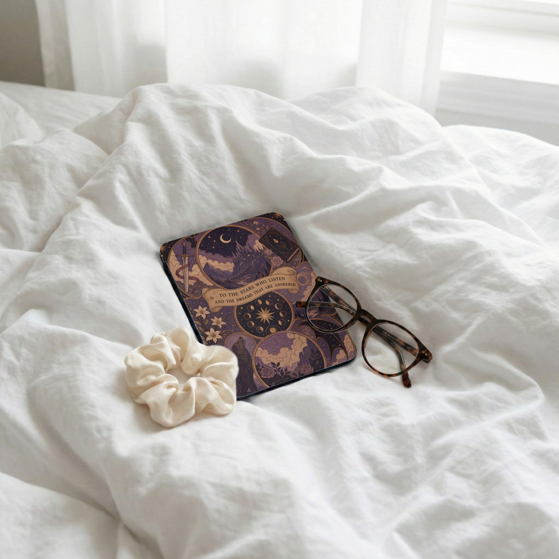 Book with celestial design and quote, scrunchie, and glasses on white bedding.