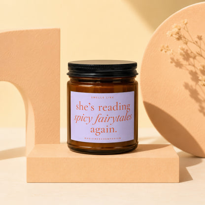 Amber glass candle with a black lid and white label that reads "she's reading spicy fairytales again."