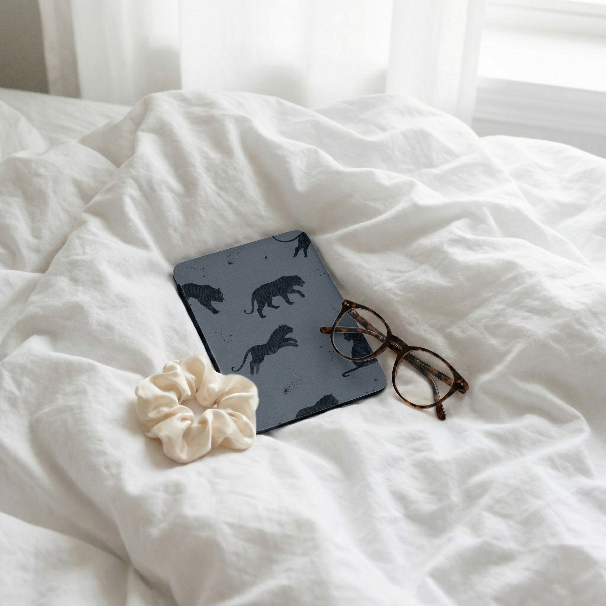 Notebook with tiger pattern, glasses, and scrunchie on white rumpled bedding with window light.