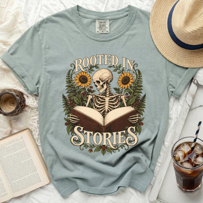 Skeleton reading a book with sunflowers and text "ROOTED IN STORIES" on t-shirt.