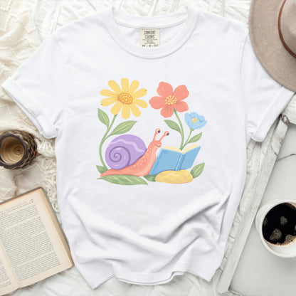 White t-shirt with a cartoon snail reading a book among flowers.