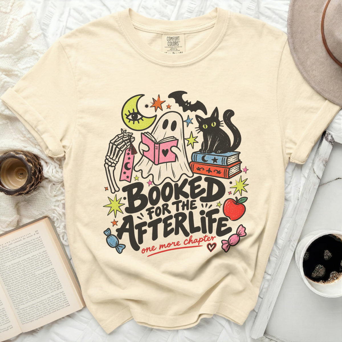 Cream t-shirt with ghost, skeleton, black cat, and books graphic. Text reads "Booked for the Afterlife, one more chapter."