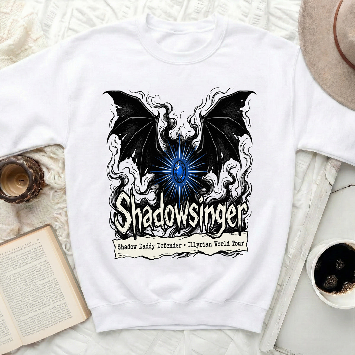 A white crewneck sweatshirt displays a black graphic featuring bat wings, a blue gem, and the text Shadowsinger and Shadow Daddy Defender - Illyrian World Tour.