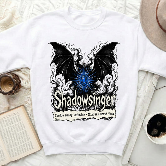 A white crewneck sweatshirt displays a black graphic featuring bat wings, a blue gem, and the text Shadowsinger and Shadow Daddy Defender - Illyrian World Tour.