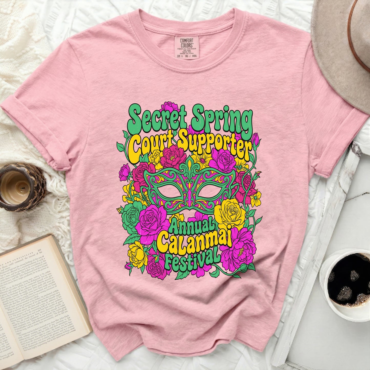 Pink t-shirt with green, yellow, and pink graphic of a mask and flowers.