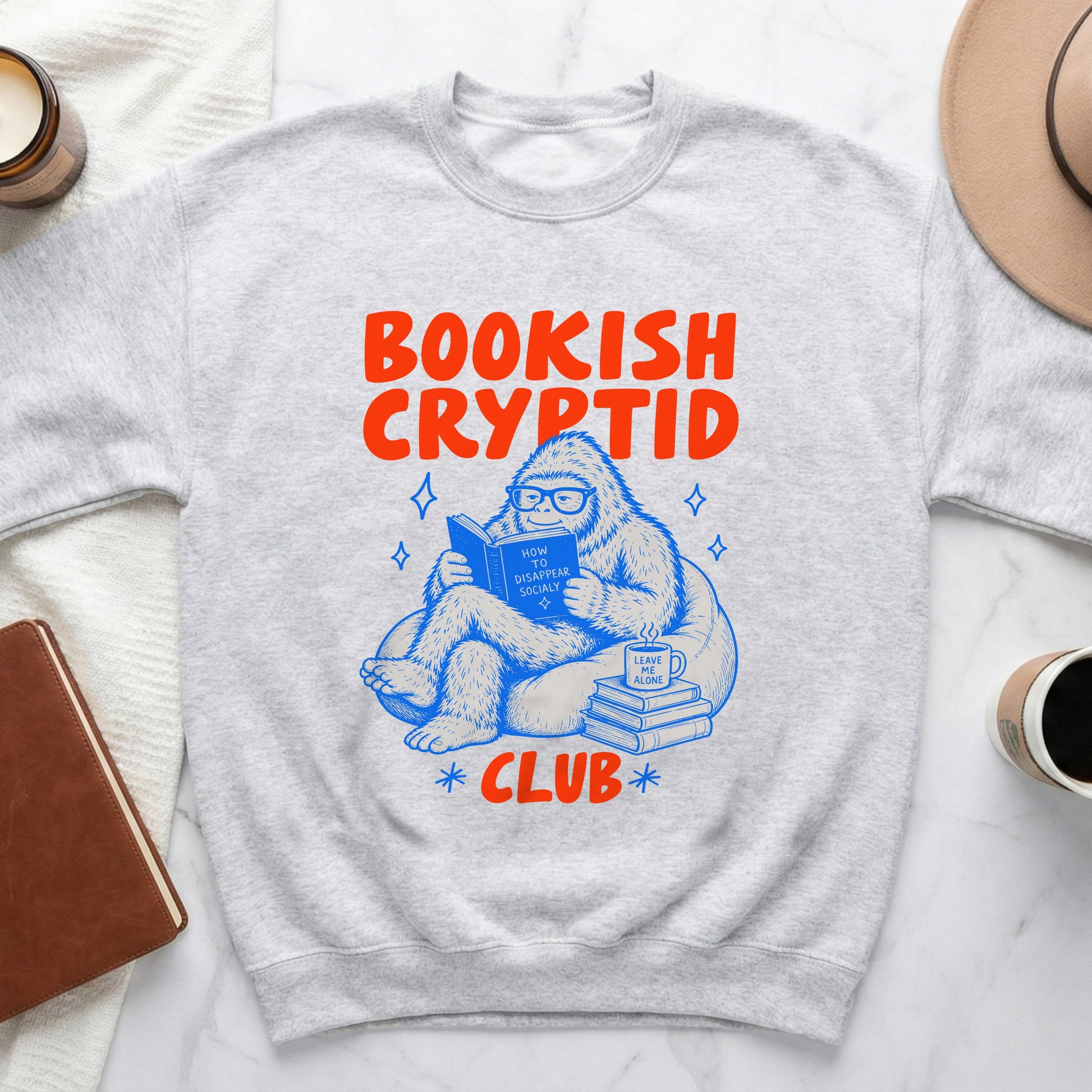 Grey sweatshirt with "Bookish Cryptid Club" text and illustration of Bigfoot reading.