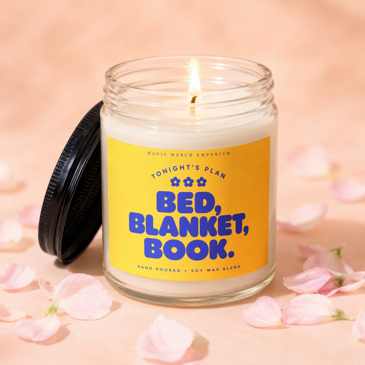 A lit soy wax candle in a clear glass jar with a yellow label reading BED, BLANKET, BOOK., surrounded by scattered pink flower petals.