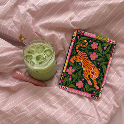 Green drink with ice, pink lip balm, and a tiger-themed notebook on pink striped fabric.
