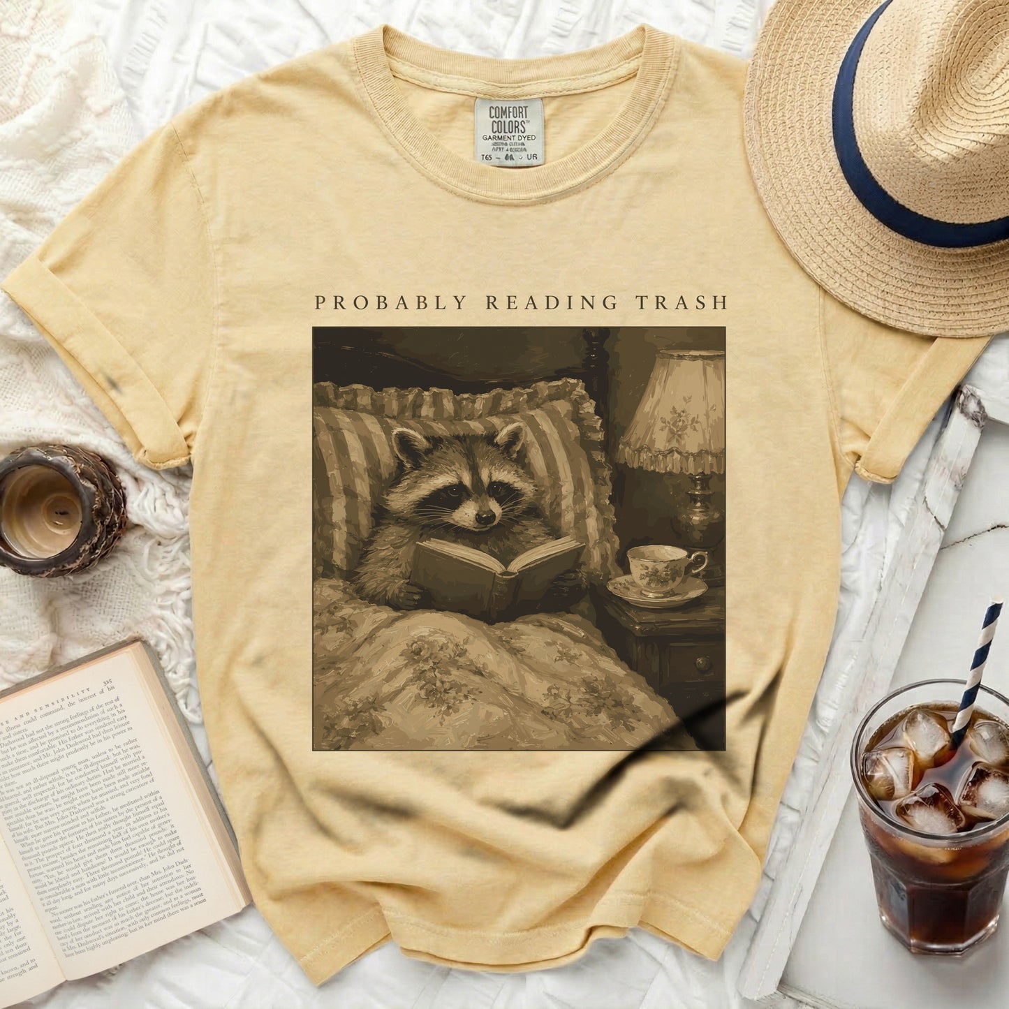 T-shirt with a graphic of a raccoon reading in bed and the text "PROBABLY READING TRASH."