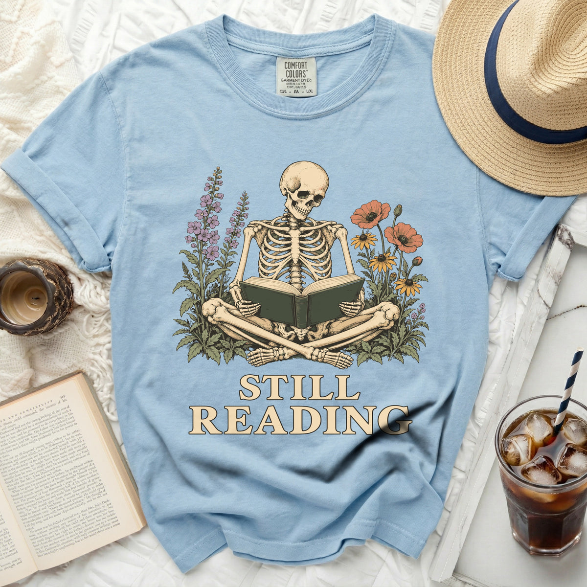 Light blue t-shirt with skeleton reading a book and "STILL READING" text.