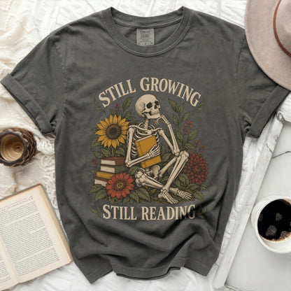 Grey t-shirt with skeleton, books, and flowers, reads "Still Growing Still Reading".