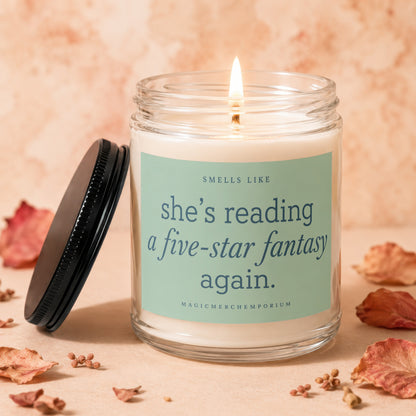 Glass candle with lit wick, reads "Smells like she's reading a five-star fantasy again."