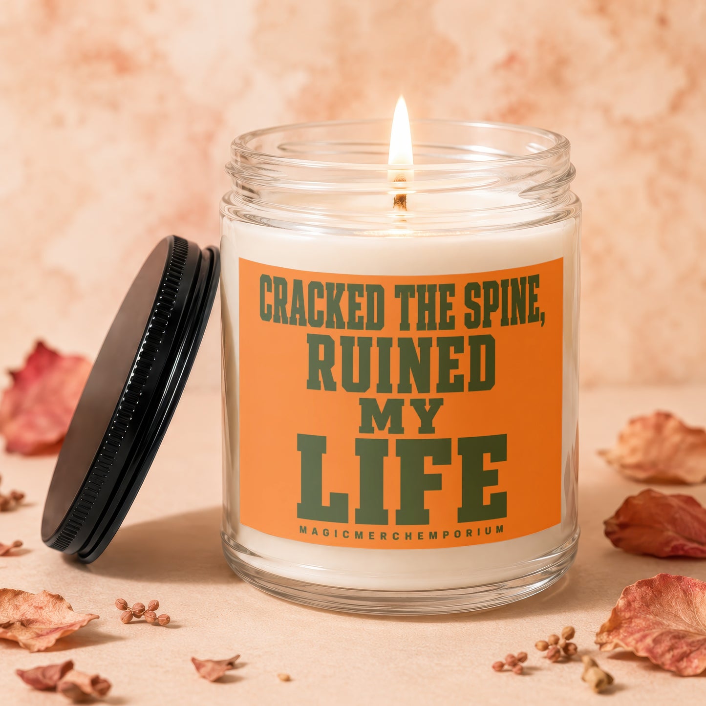 Glass candle with lit wick and orange label reading "CRACKED THE SPINE, RUINED MY LIFE".