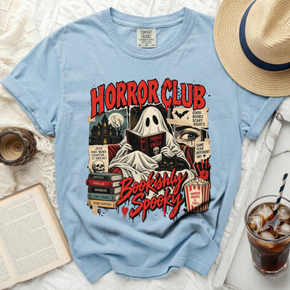 Light blue t-shirt featuring a spooky graphic of a ghost reading a book, accompanied by stacked books, a black cat, and red text that reads Horror Club Bookishly Spooky.