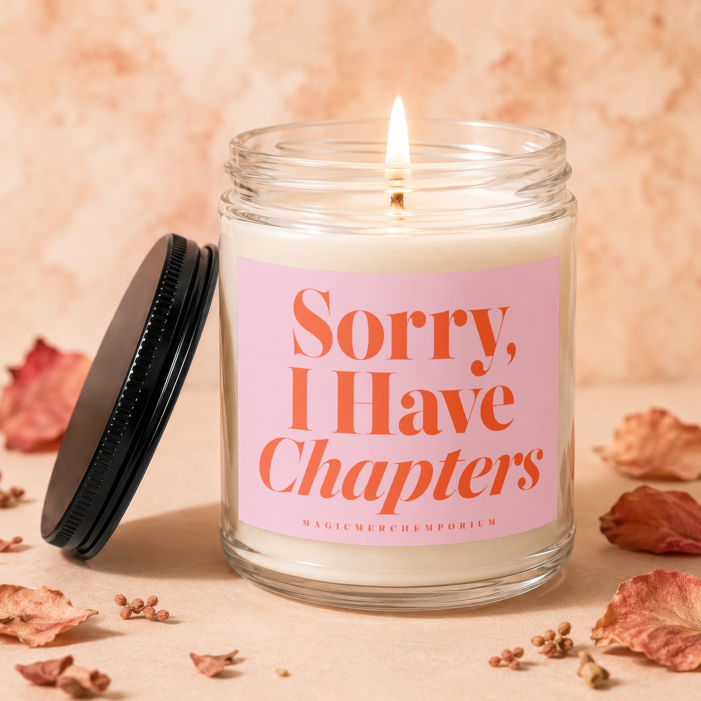 Glass candle with lit wick and pink label reading "Sorry, I Have Chapters"