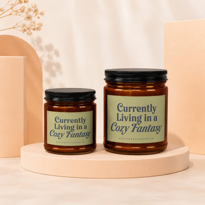 Two amber glass candles with black lids, labeled "Currently Living in a Cozy Fantasy".
