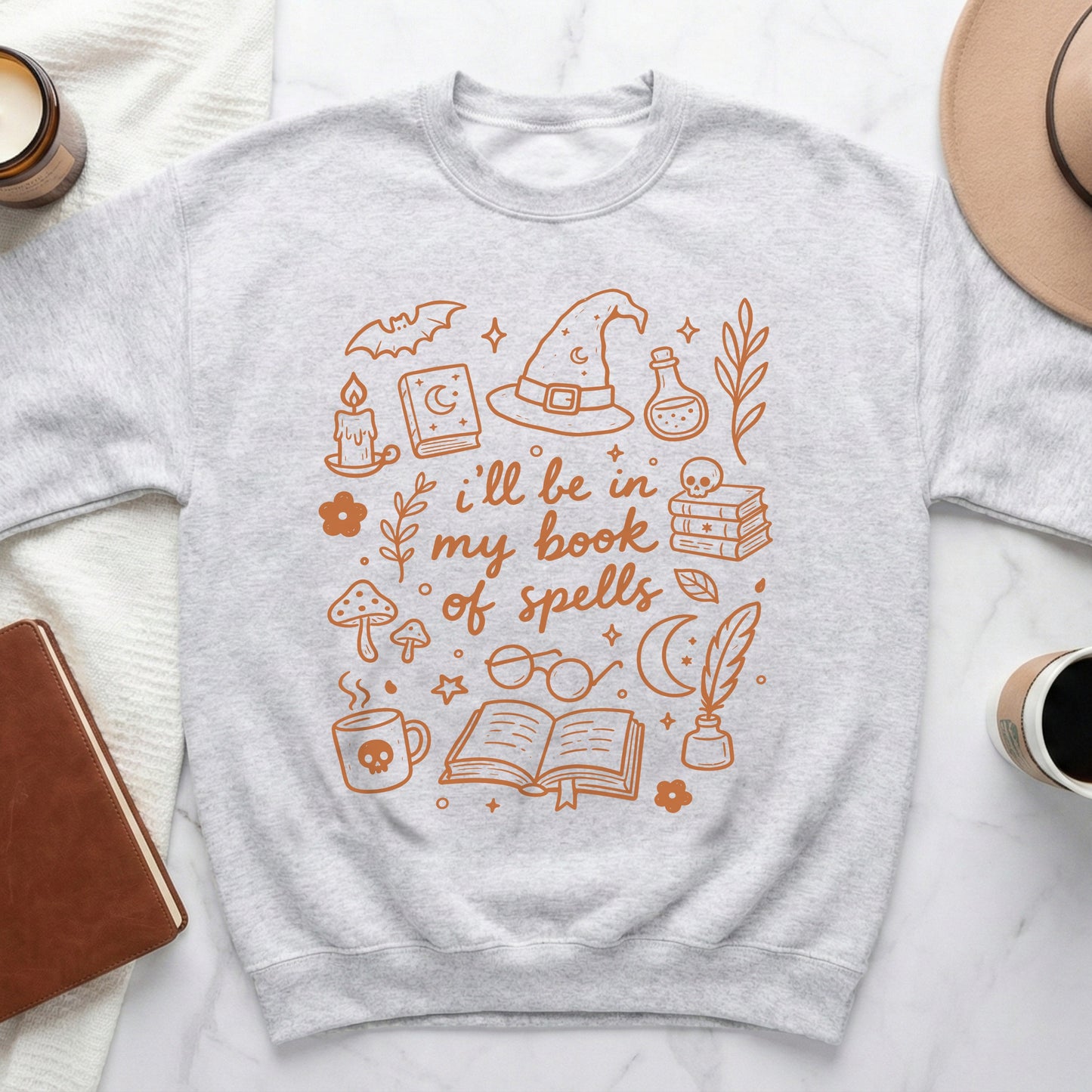 Light grey sweatshirt with orange line art of witchy items and text "i'll be in my book of spells".
