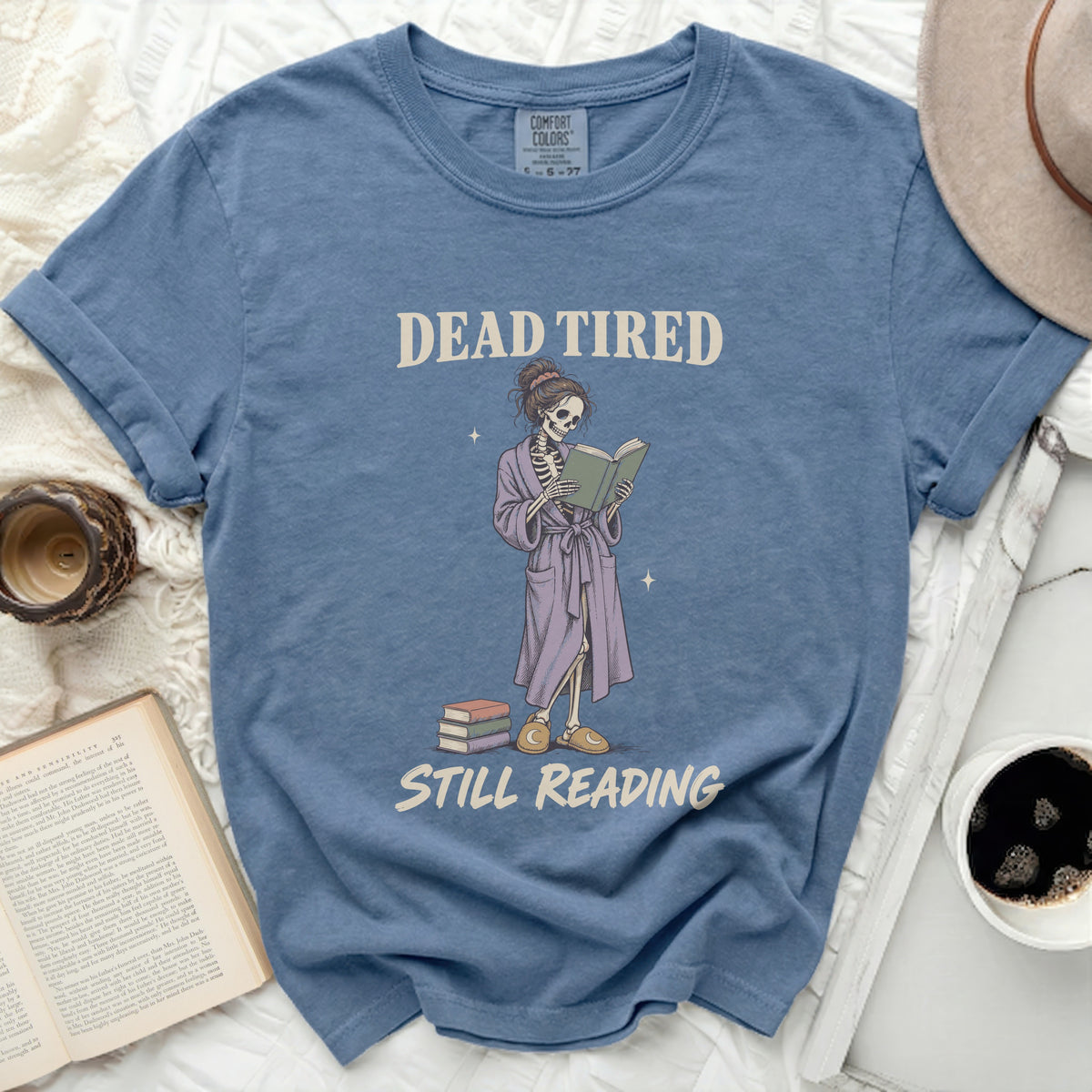 Skeleton wearing bathrobe reads a book with text "DEAD TIRED STILL READING".