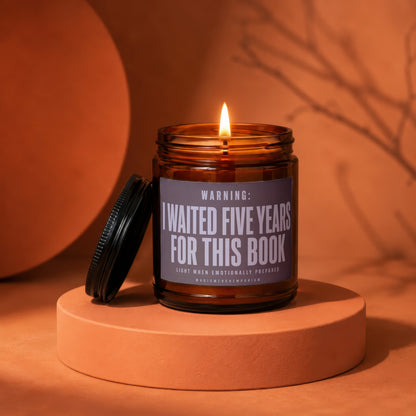 A lit amber glass jar candle with a black lid rests on an orange pedestal, featuring a label that reads Warning: I Waited Five Years For This Book.