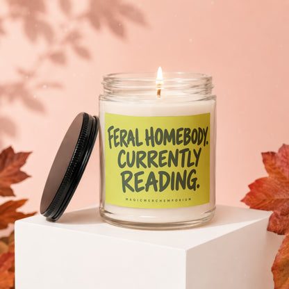 Glass candle with lit wick and black lid, reads "Feral Homebody Currently Reading."