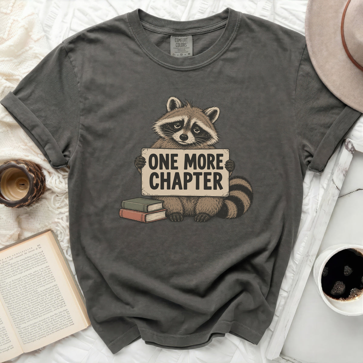 Raccoon holding "One More Chapter" sign with books and coffee nearby.
